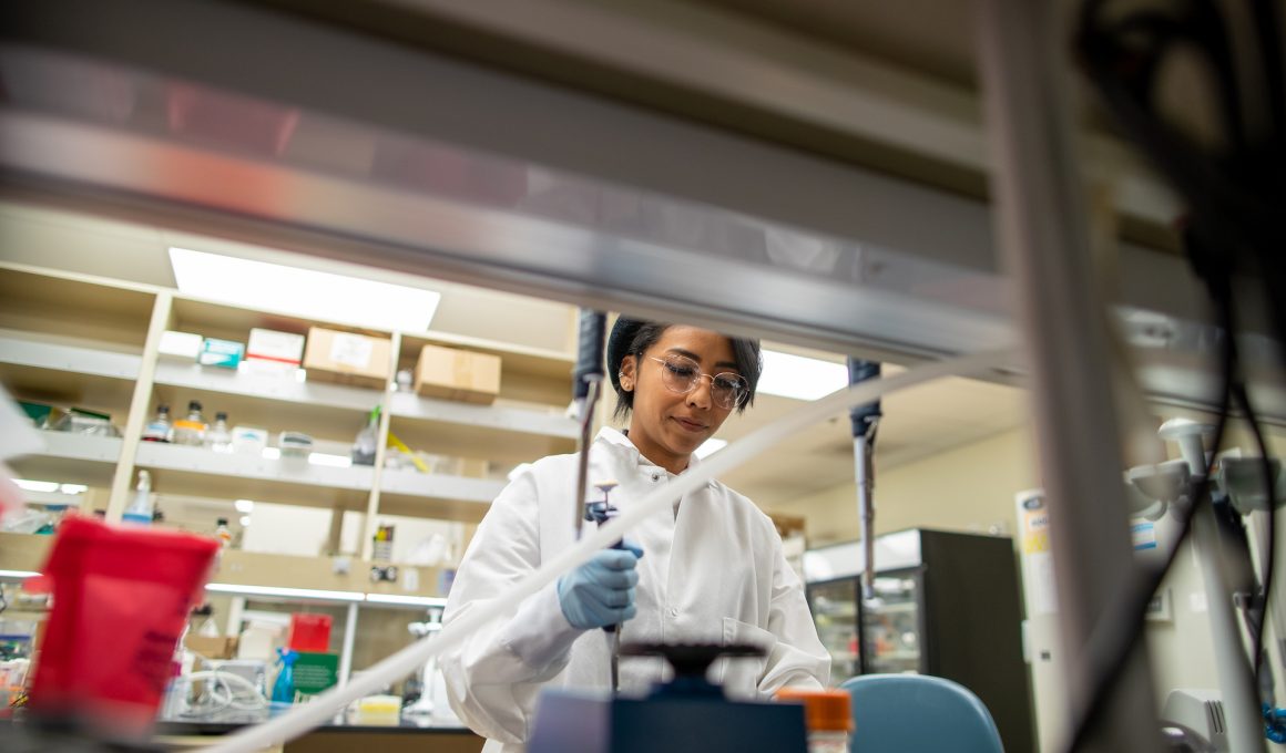 Photo of a scientists in the laboratory of Samuel Myers, Ph.D. We see the scientists through a window in shelving. They are wearing a white coat and using a pipette