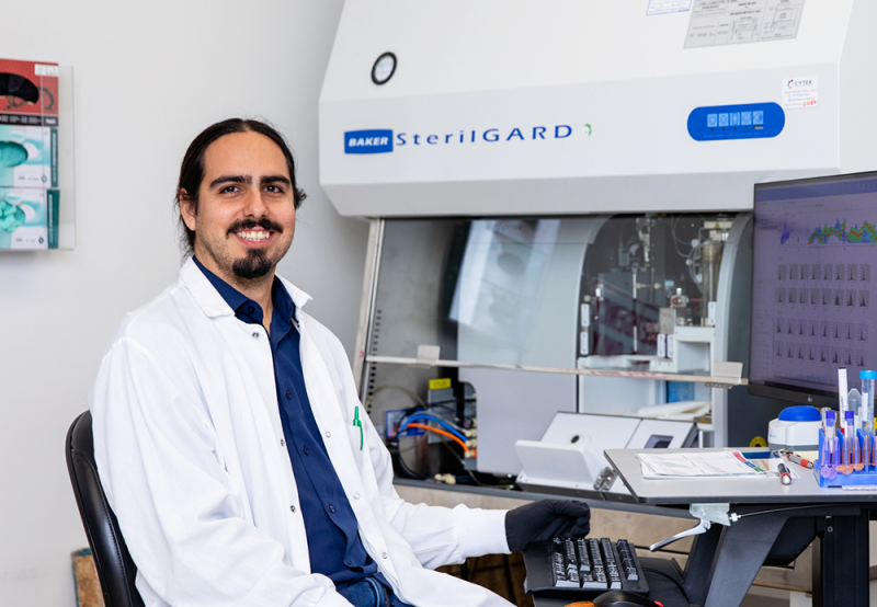 Photo of LJI scientist Gabriel Ascui Gac. He is wearing gloves and a white lab coat. Sitting in front of a computer in a laboratory space with a sterile hood