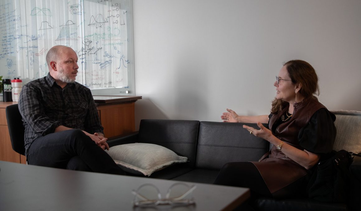 Sam Myers sits in an office chair while speaking with Sandra Boek Werness.