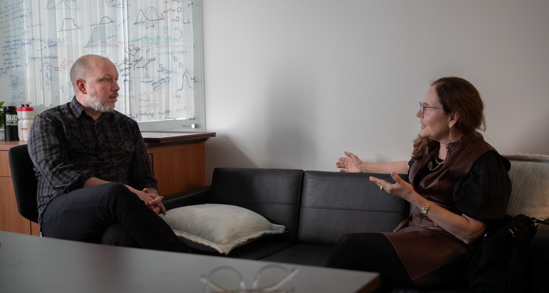 Sam Myers sits in an office chair while speaking with Sandra Boek Werness.