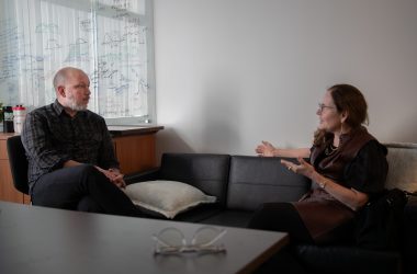 Sam Myers sits in an office chair while speaking with Sandra Boek Werness.