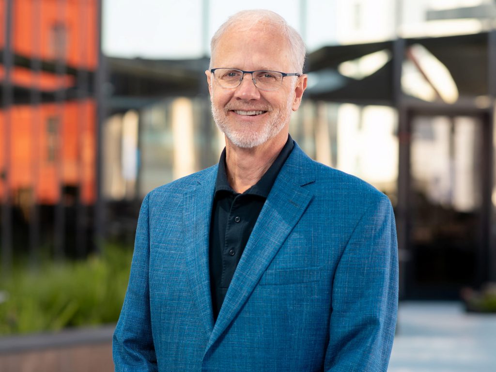 A man in a blue suit stands outside. He is smiling at the camera. He has white hair and glasses