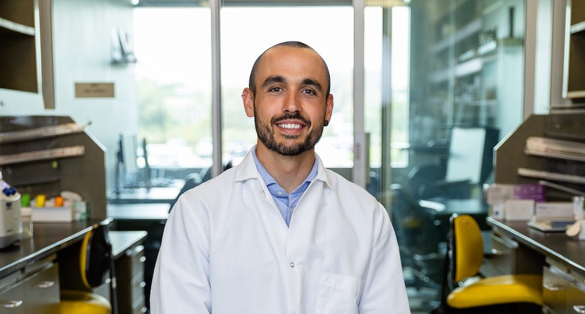Dr. Reina sits in his laboratory. He is wearing a white lab coat and looking at the camera