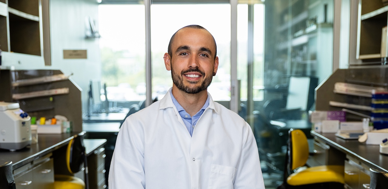 Dr. Reina sits in his laboratory. He is wearing a white lab coat and looking at the camera