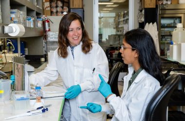 Daniela Weiskopf (left) in the lab with Rimjhim Agarwal. They are at the lab bench, wearing white lab coats and gloves. They are looking at each other and talking